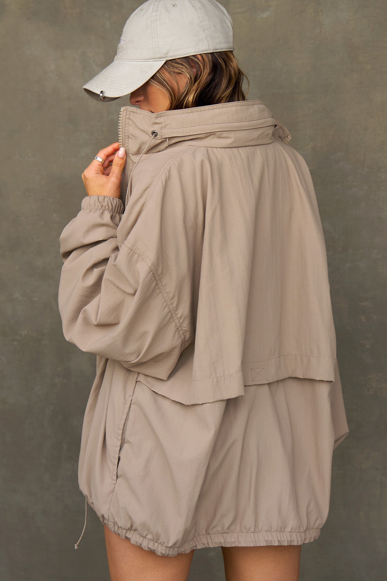 Back view of model posing in the oversized fawn nylon Classic Windbreaker Jacket with a collar, zip-away hood and reflective corded details at the hem, sleeves and neck for cinching.