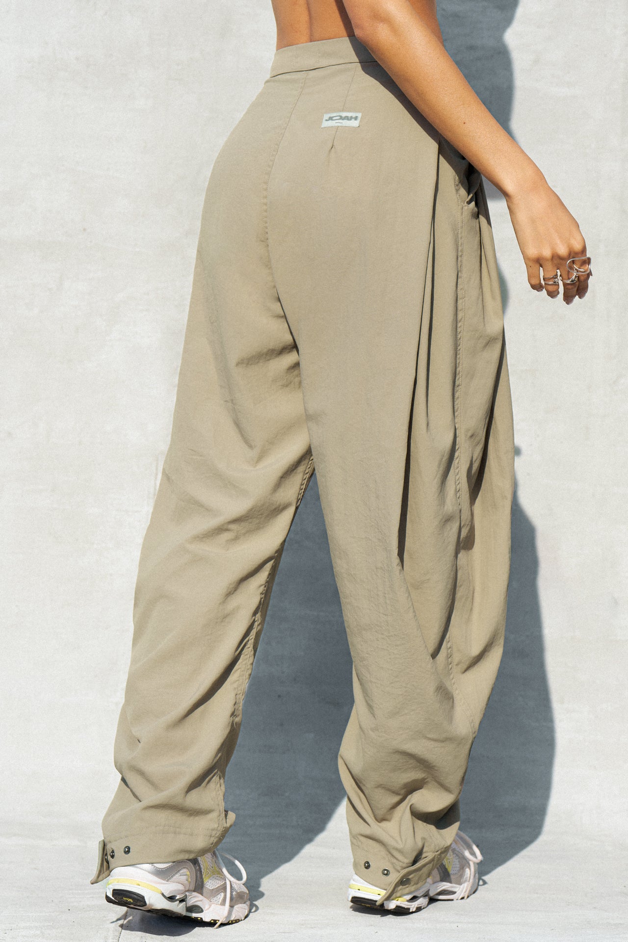 Back view of model from the waist down posing in the oversized, horseshoe-style desert sage nylon Barrel Pant with side pockets, zip fly, pleated sides, adjustable snap closures at the ankle and a JOAH BROWN logo on the back below the waistline.
