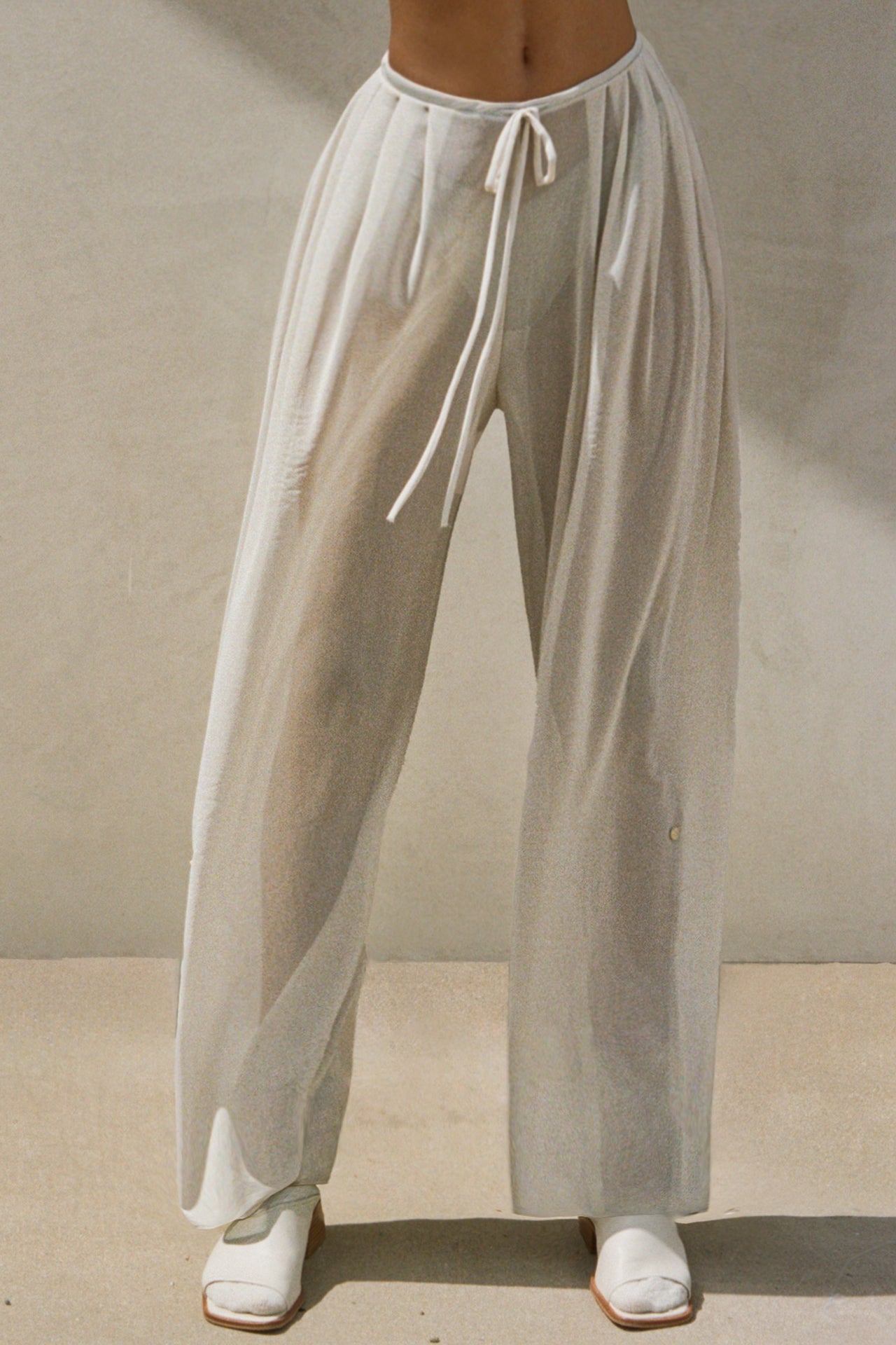 Front view of model from the waist down posing in the relaxed fit porcelain mesh Pleated Sheer Pant with a wide leg, elastic waistband, adjustable drawstring tie and buttons on the inner and outer leg for cinching.
