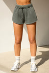 Front view of model from the waist down posing in the relaxed fit washed sage french terry Sweat Short with side pockets, hidden drawstring, adjustable elastic waistband and a Joah Brown logo patch at the bottom side of the left leg.