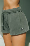 Close up detail side view of model from the waist down posing in the relaxed fit washed sage french terry Sweat Short with side pockets, hidden drawstring, adjustable elastic waistband and a Joah Brown logo patch at the bottom side of the left leg.