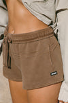 Close up detail side view of model from the waist down posing in the relaxed fit cocoa french terry Sweat Short with side pockets, hidden drawstring,adjustable elastic waistband and a Joah Brown logo patch at the bottom side of the left leg.