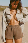 Front view of model posing in the relaxed fit cocoa french terry Sweat Short with side pockets, hidden drawstring,adjustable elastic waistband and a Joah Brown logo patch at the bottom side of the left leg.