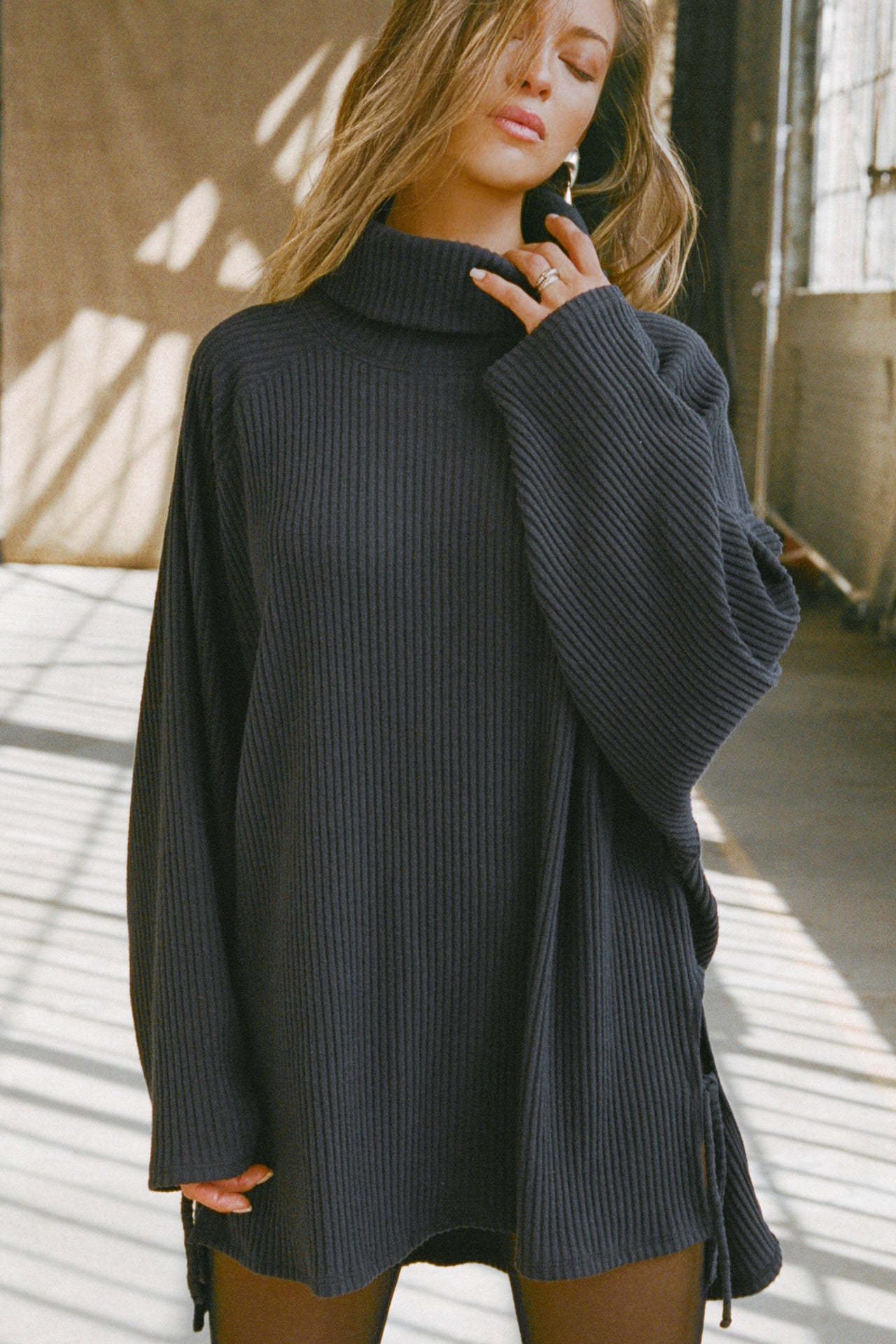 Front view of model posing in the relaxed and slouchy tunic-style midnight luxe knit Turtleneck Tunic with dropped shoulders, side slits and adjustable ties for a customized shape