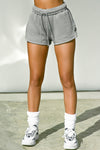 Front view of model from the waist down posing in the relaxed fit stone french terry Sweat Short with side pockets, hidden drawstring, adjustable elastic waistband and a Joah Brown logo patch at the bottom side of the left leg.