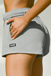 Close up detail side view of model from the waist down posing in the relaxed fit stone french terry Sweat Short with side pockets, hidden drawstring, adjustable elastic waistband and a Joah Brown logo patch at the bottom side of the left leg.