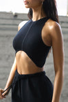 Side view of model posing in the fitted cropped black flexrib Curve Crop Tank wih a vertical stitch detail down the front, a curved hemline in an inverted u-shape and a high v-neckline