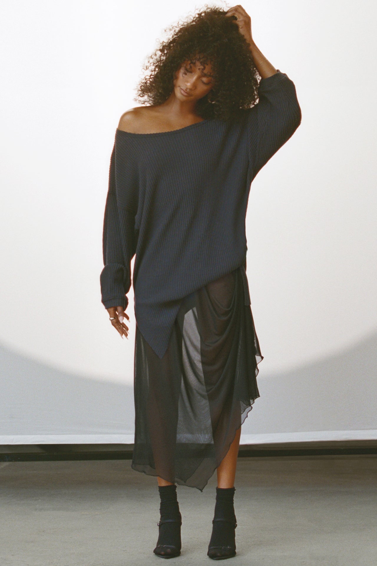 Full body front view of model posing in the lightweight black mesh Asymmetrical Button Skirt with a draped silhouette, asymmetrical hemline and functional side button details for an adjustable fit