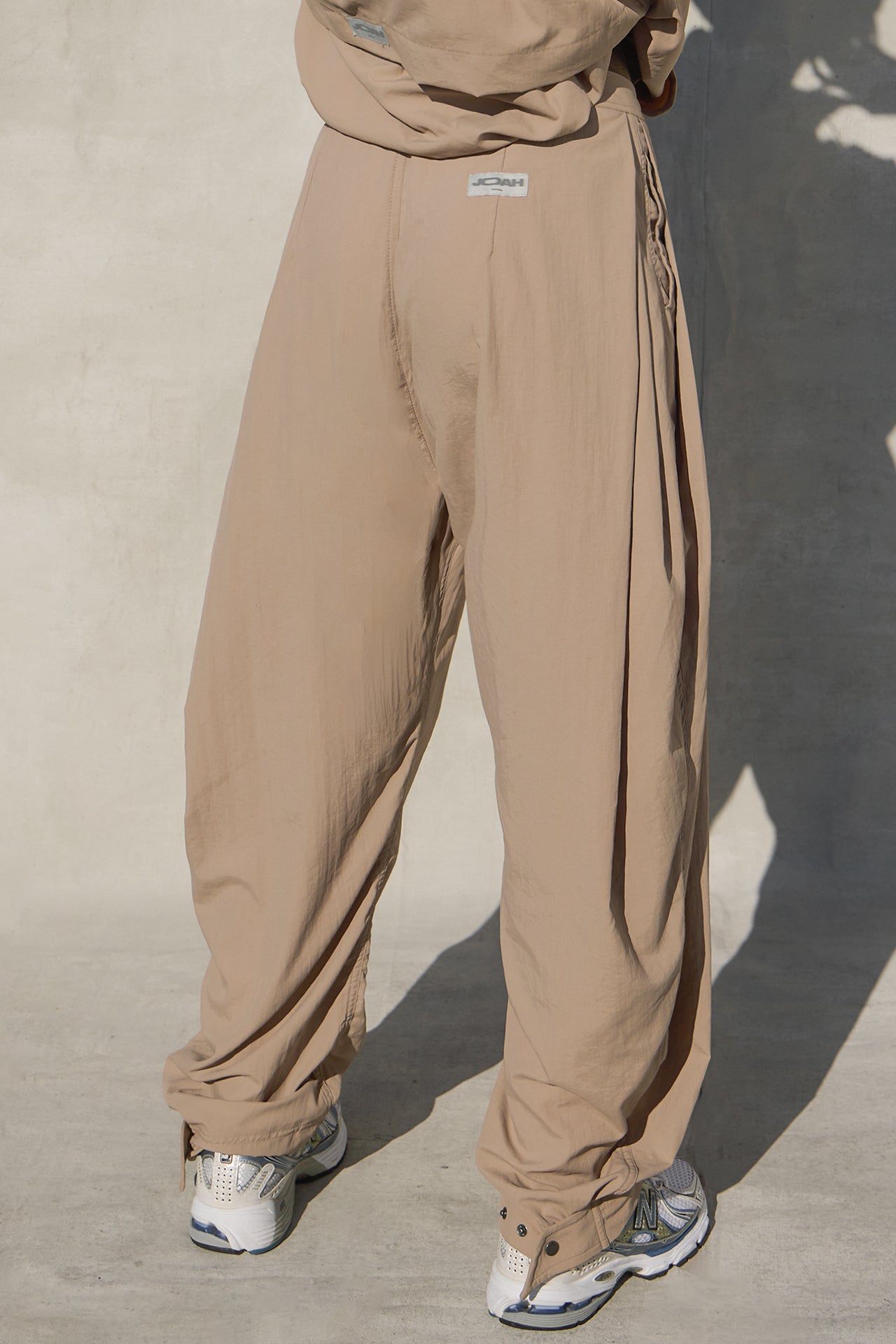 Back view of model from the waist down posing in the oversized, horseshoe-style fawn nylon Barrel Pant with side pockets, zip fly, pleated sides, adjustable snap closures at the ankle and a JOAH BROWN logo on the back below the waistline.