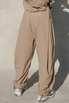 Back view of model from the waist down posing in the oversized, horseshoe-style fawn nylon Barrel Pant with side pockets, zip fly, pleated sides, adjustable snap closures at the ankle and a JOAH BROWN logo on the back below the waistline.