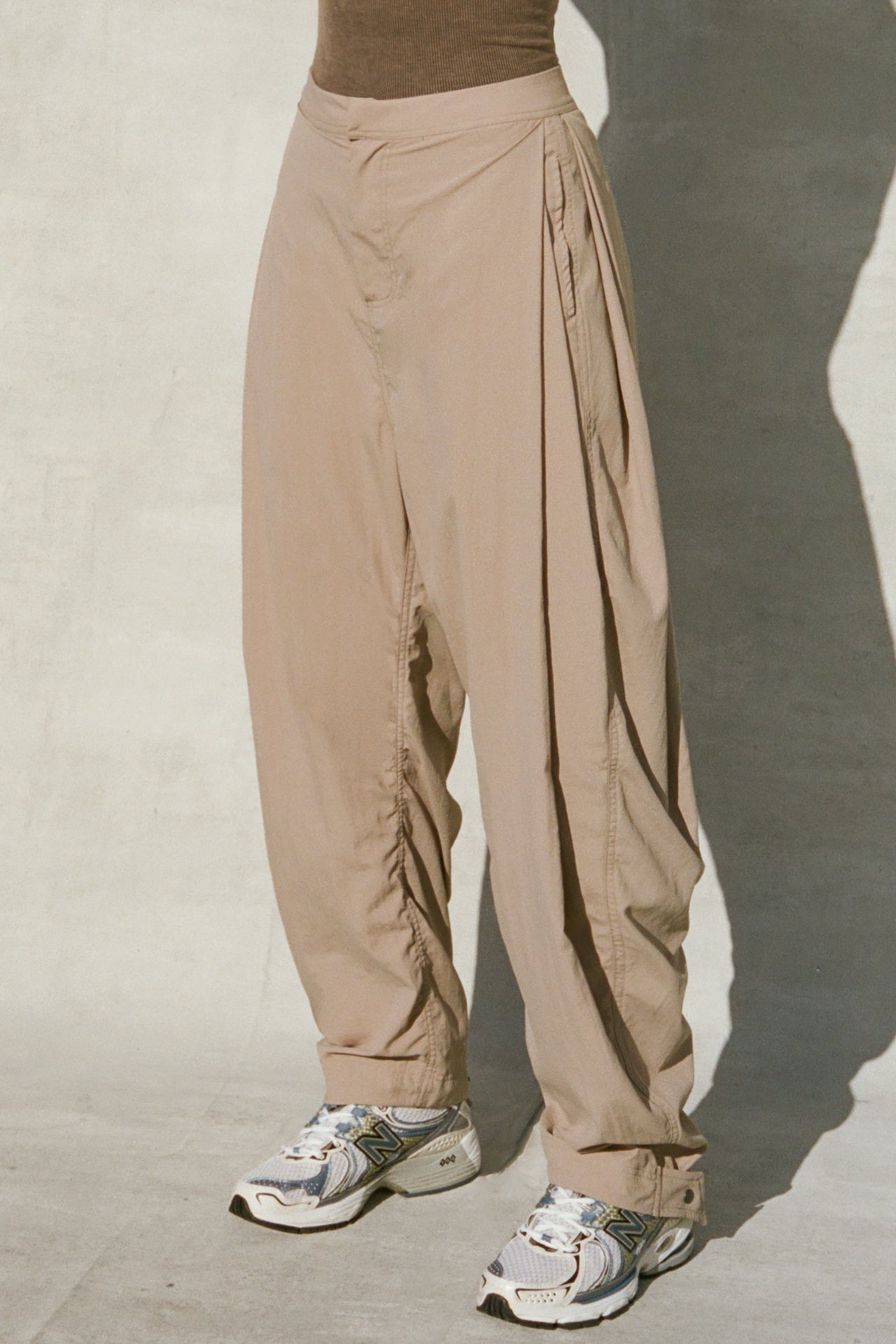 Front view of model from the waist down posing in the oversized, horseshoe-style fawn nylon Barrel Pant with side pockets, zip fly, pleated sides and adjustable snap closures at the ankle.