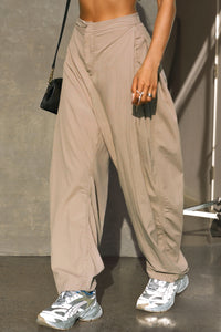 Front view of model from the waist down posing in the oversized, horseshoe-style fawn nylon Barrel Pant with side pockets, zip fly, pleated sides and adjustable snap closures at the ankle.