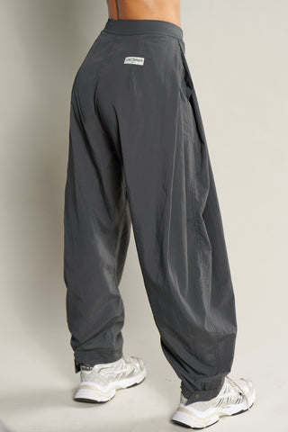Back view of model from the waist down posing in the oversized, horseshoe-style jet nylon Barrel Pant with side pockets, zip fly, pleated sides, adjustable snap closures at the ankle and a JOAH BROWN logo on the back below the waistline.