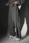 Side view of model from the waist down posing in the oversized, horseshoe-style jet nylon Barrel Pant with side pockets, zip fly, pleated sides and adjustable snap closures at the ankle.