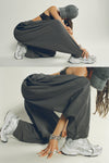 Two side views of model stretching and posing in the oversized, horseshoe-style jet nylon Barrel Pant with side pockets, zip fly, pleated sides and adjustable snap closures at the ankle.