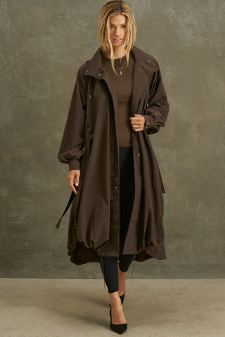Full body front view of model posing in the espresso nylon City Trench coat with oversized silhouette, structured collar, attached hood, self-tie belt and adjustable sweep