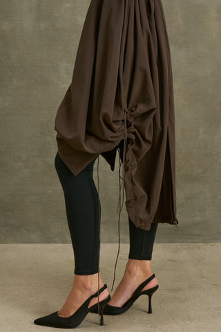 Side view of model from the waist down posing in the espresso nylon City Trench coat with oversized silhouette, structured collar, attached hood, self-tie belt and adjustable sweep