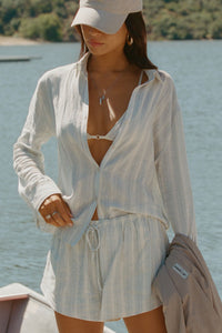 Front view of model posing outside by a lake in the lightweight natural white and blue stripe gauze Classic Button Down collared long sleeve shirt with front button closures