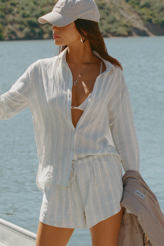 Front view of model posing outside by a lake in the lightweight natural white and blue stripe gauze Classic Button Down collared long sleeve shirt with front button closures
