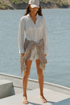 Full body front view of model posing outside by a lake in the lightweight natural white and blue stripe gauze Classic Button Down collared long sleeve shirt with front button closures