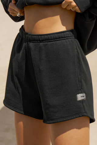 Close up side view of model from the waist down posing in the relaxed fit black french terry Classic Sweat Short with side pockets, hidden drawstring, adjustable elastic waistband and JOAH BROWN logo patch on the bottom left leg