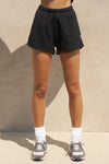 Front view of model from the waist down posing in the relaxed fit black french terry Classic Sweat Short with side pockets, hidden drawstring, adjustable elastic waistband and JOAH BROWN logo patch on the bottom left leg