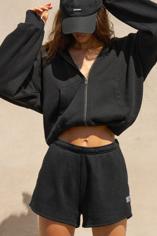 Front view of model posing in the relaxed fit black french terry Classic Sweat Short with side pockets, hidden drawstring, adjustable elastic waistband and JOAH BROWN logo patch on the bottom left leg