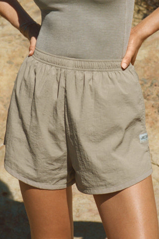 Close up front view of model from the waist down posing outside in the relaxed-fit desert sage nylon Classic Track Short with an elastic waistband, side seam pockets, and adjustable drawcord.