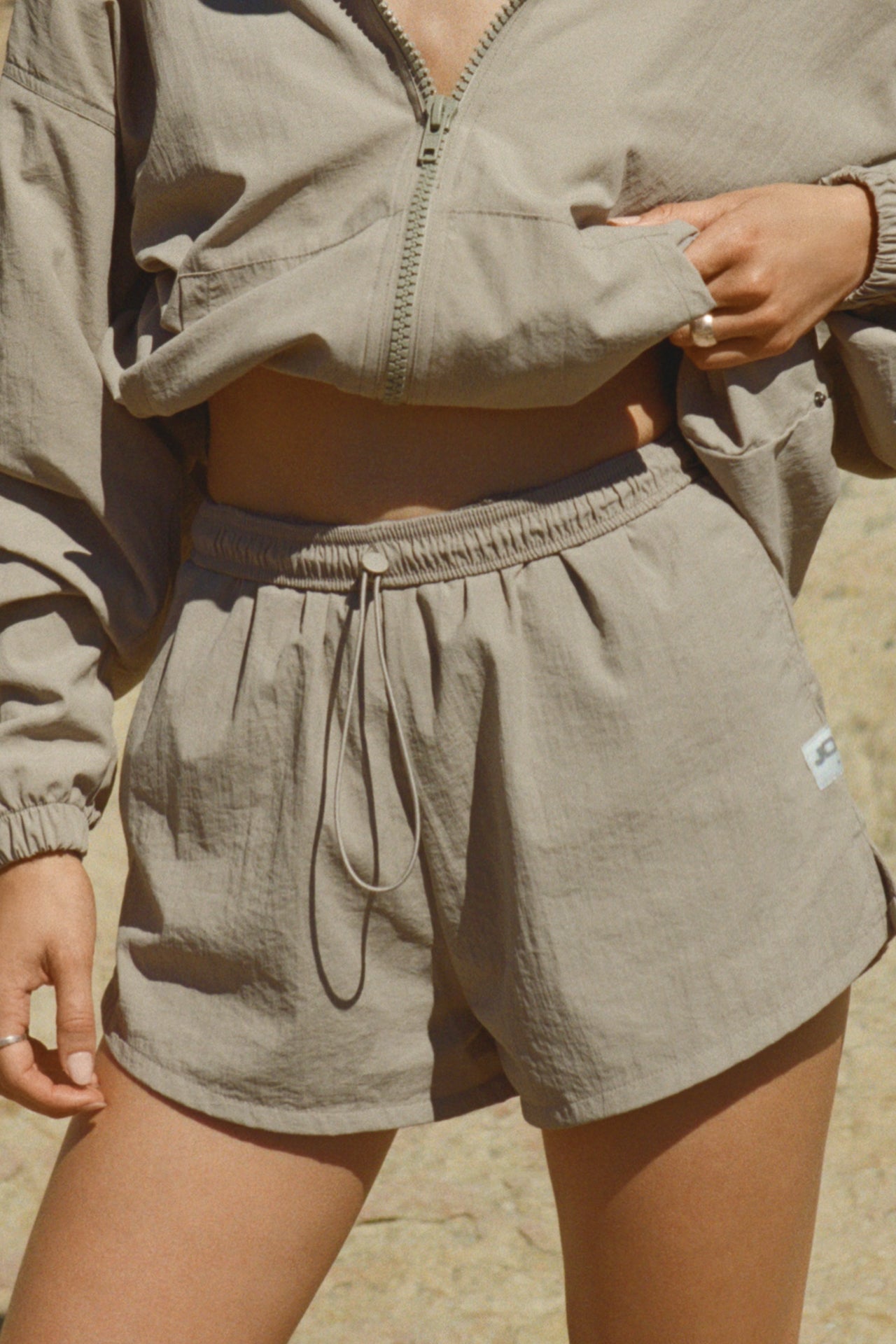 Close up front view of model from the waist down posing outside in the relaxed-fit desert sage nylon Classic Track Short with an elastic waistband, side seam pockets, and adjustable drawcord.