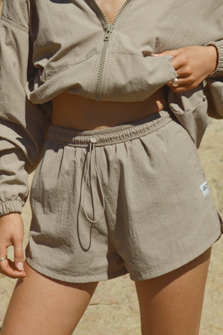 Close up front view of model from the waist down posing outside in the relaxed-fit desert sage nylon Classic Track Short with an elastic waistband, side seam pockets, and adjustable drawcord.