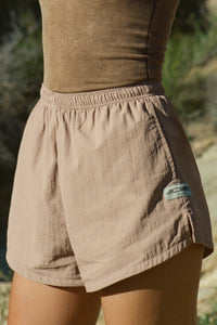 Close up side view of model from the waist down posing outside in the relaxed-fit fawn nylon Classic Track Short with an elastic waistband, side seam pockets, and adjustable drawcord.