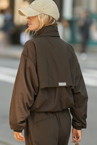 Back view of model posing in a city  in the relaxed-fit espresso nylon Classic Windbreaker Jacket with a padded collared neck for structure & adjustable corded details at the sweep for cinching