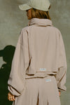 Back view of model posing in the oversized fawn nylon Classic Windbreaker Jacket with a collar, zip-away hood and reflective corded details at the hem, sleeves and neck for cinching.