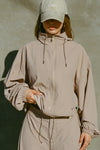 Front view of model posing in the oversized fawn nylon Classic Windbreaker Jacket with a collar, zip-away hood and reflective corded details at the hem, sleeves and neck for cinching.