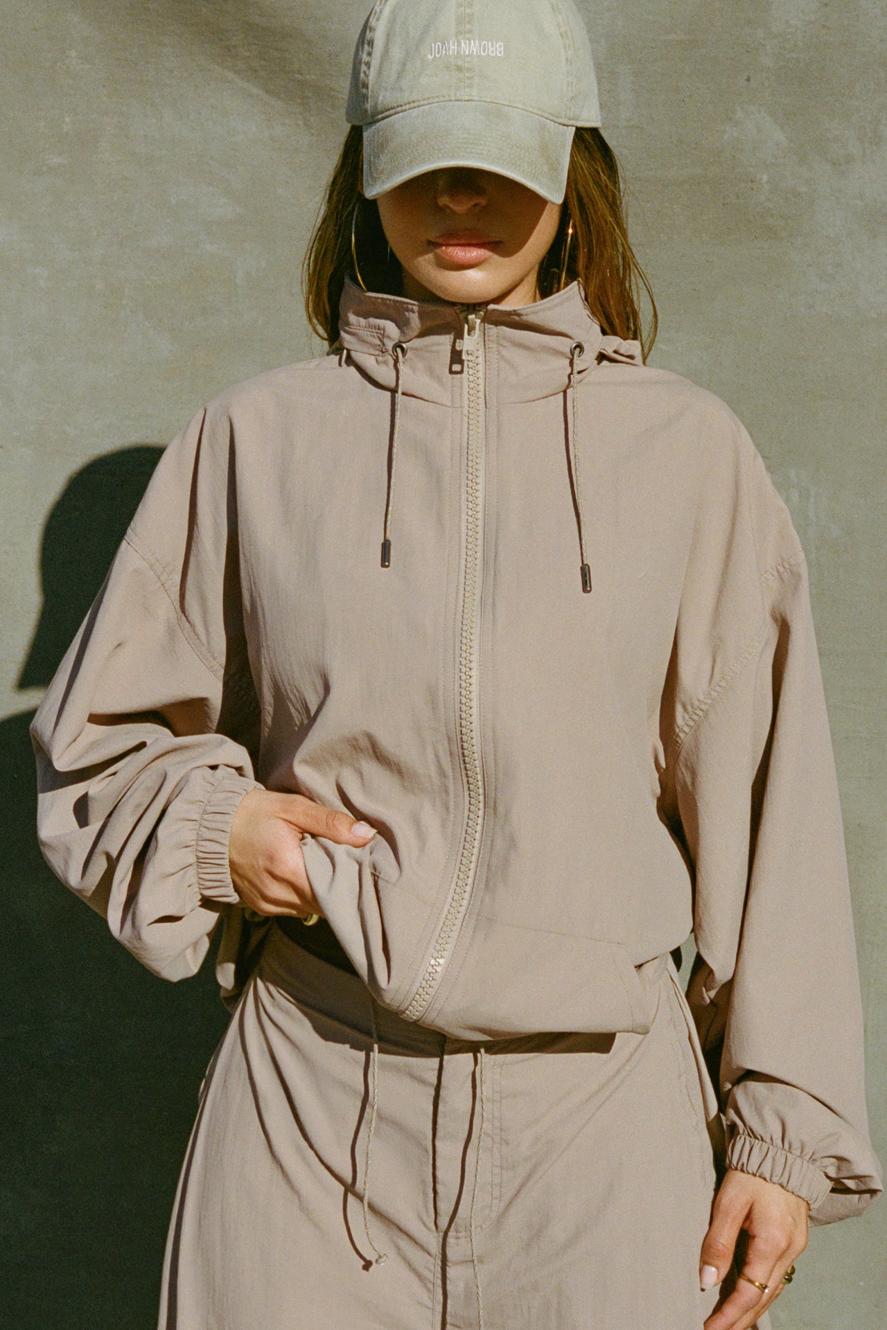 Front view of model posing in the oversized fawn nylon Classic Windbreaker Jacket with a collar, zip-away hood and reflective corded details at the hem, sleeves and neck for cinching.