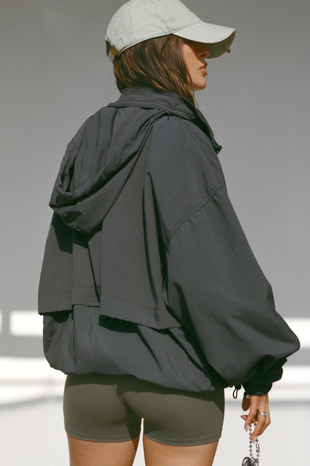 Back view of model posing in the oversized jet nylon Classic Windbreaker Jacket with a collar, zip-away hood and reflective corded details at the hem, sleeves and neck for cinching.