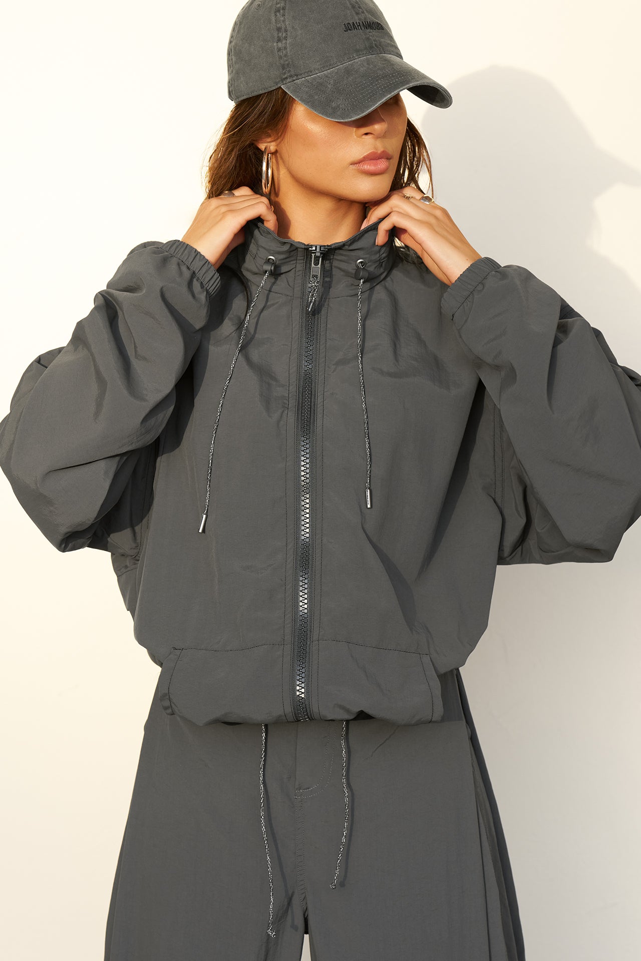 Front view of model posing in the oversized jet nylon Classic Windbreaker Jacket with a collar, zip-away hood and reflective corded details at the hem, sleeves and neck for cinching.