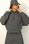Front view of model posing in the oversized jet nylon Classic Windbreaker Jacket with a collar, zip-away hood and reflective corded details at the hem, sleeves and neck for cinching.