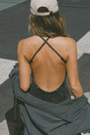 Back view of model posing outside in the street in the fitted  charcoal washed rib Cross Back Midi Dress with an asymmetrical hem, a low scoop back and thin adjustable straps that criss cross in the back