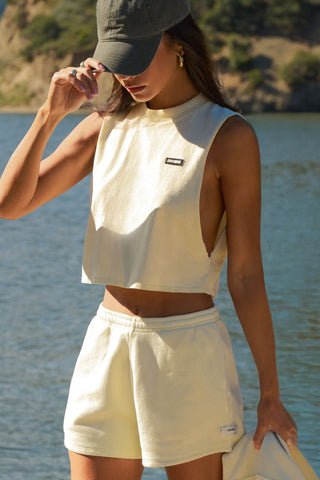 Front view of model posing outside by a lake in the relaxed fit buttercream cotton Cut Off Boyfriend Tank with low cut armholes, a crew neckline and a joah brown logo patch at the upper left chest