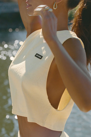 Close up side view of model posing outside by a lake in the relaxed fit buttercream cotton Cut Off Boyfriend Tank with low cut armholes, a crew neckline and a joah brown logo patch at the upper left chest
