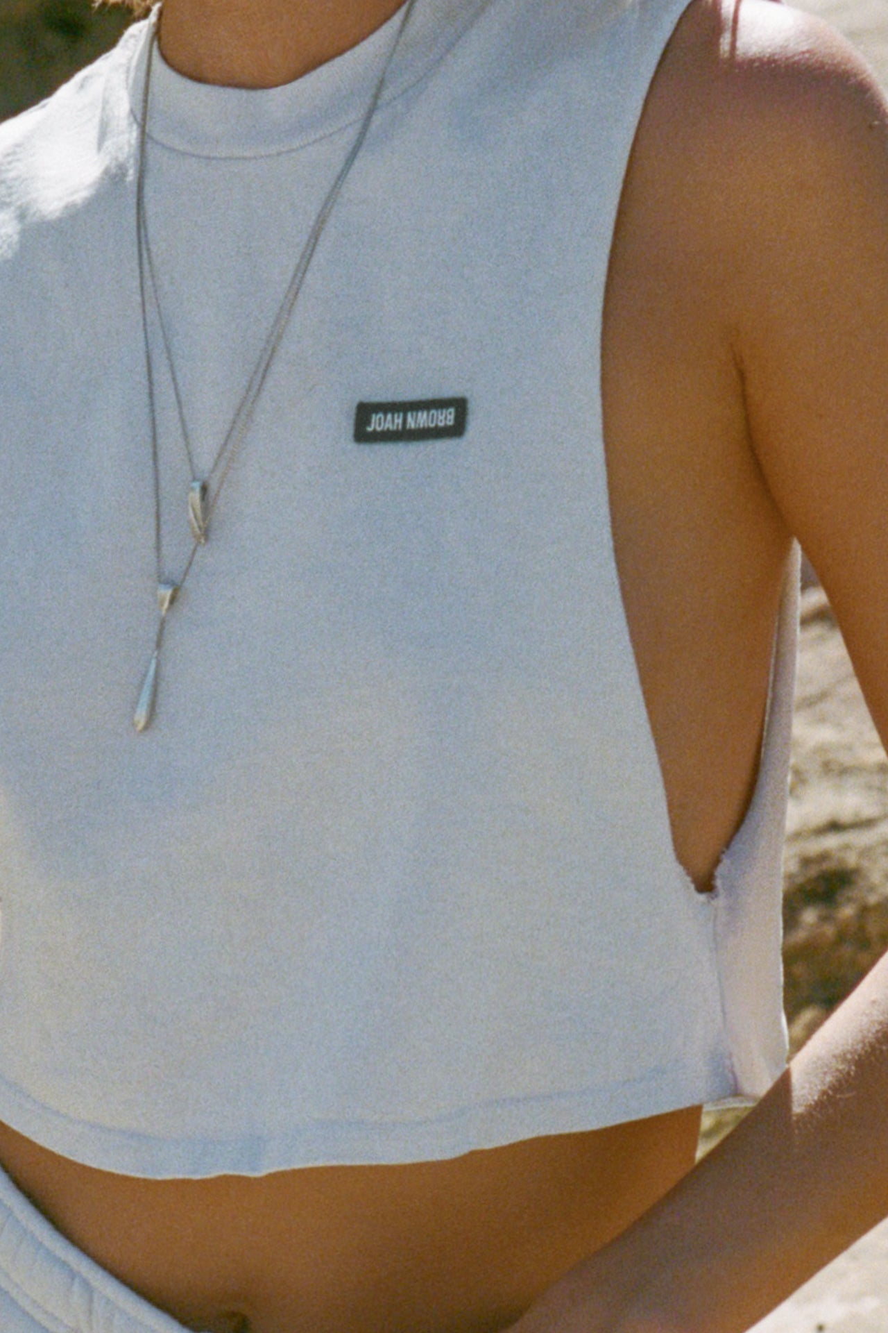Close up detail front view of model posing outside by a truck in the relaxed fit cloud cotton Cut Off Boyfriend Tank with low cut armholes, a crew neckline and a joah brown logo patch at the upper left chest