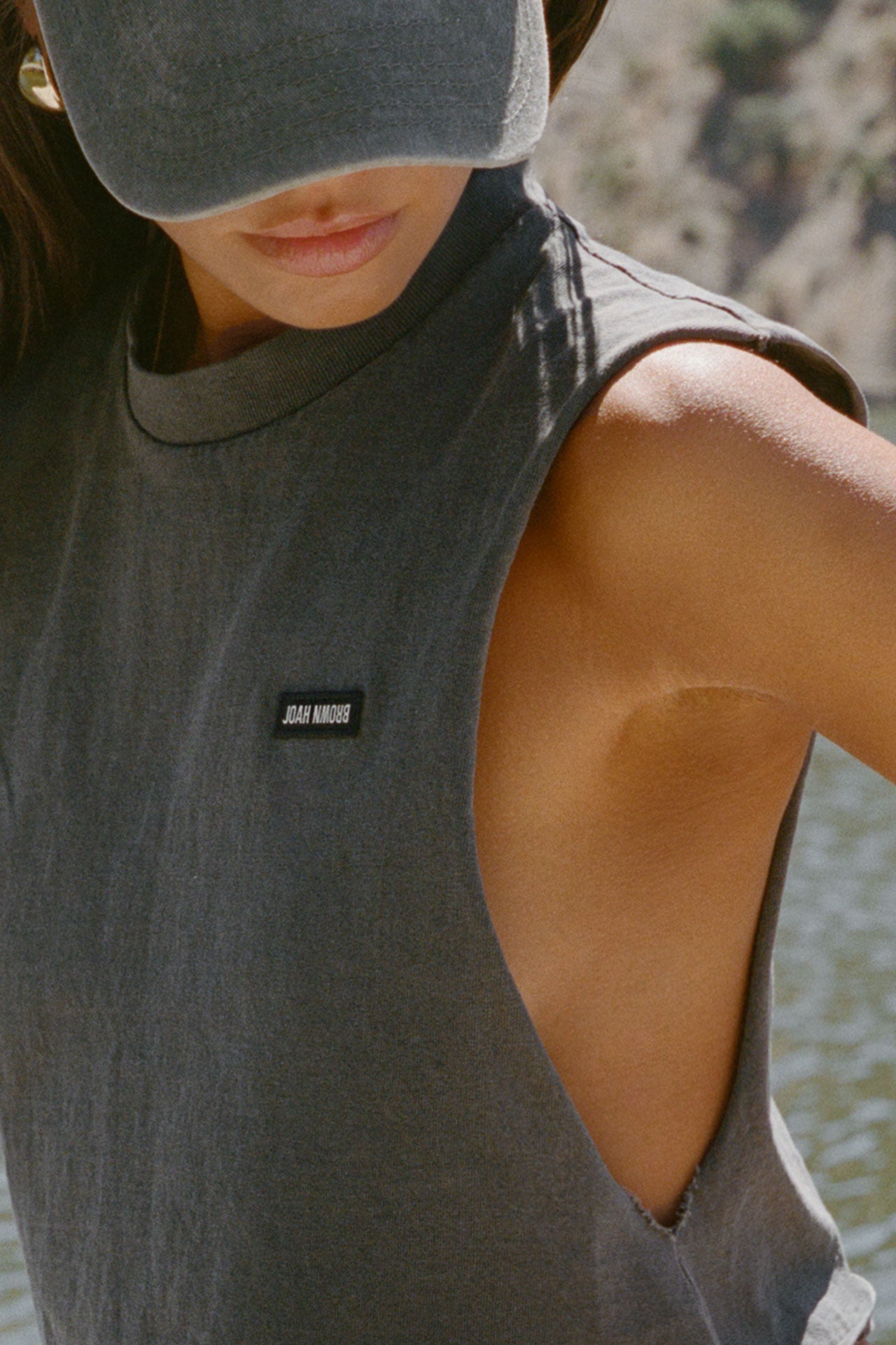 Close up detail side view of model posing outside by a lake in the relaxed fit washed black cotton Cut Off Boyfriend Tank with low cut armholes, a crew neckline and a joah brown logo patch at the upper left chest
