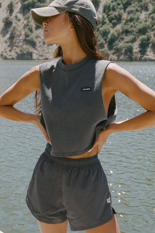 Front view of model posing outside by a lake in the relaxed fit washed black cotton Cut Off Boyfriend Tank with low cut armholes, a crew neckline and a joah brown logo patch at the upper left chest