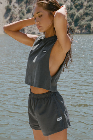 Side view of model posing outside by a lake in the relaxed fit washed black cotton Cut Off Boyfriend Tank with low cut armholes, a crew neckline and a joah brown logo patch at the upper left chest