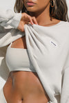 Close up detail front view of model posing in the loose fit cropped sahara french terry Cut Off Sweatshirt with oversized long sleeves, a raw cut hem and a white patch with a black joah brown logo on it sewn on the upper left chest