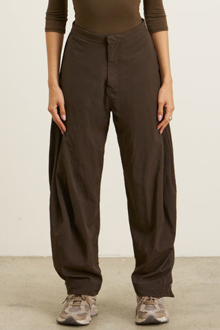 Front view of model from the waist down posing in the oversized, horseshoe-style espresso nylon Barrel Pant with side pockets, zip fly, pleated sides and adjustable snap closures at the ankle.