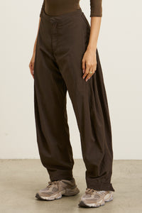 Front view of model from the waist down posing in the oversized, horseshoe-style espresso nylon Barrel Pant with side pockets, zip fly, pleated sides and adjustable snap closures at the ankle.