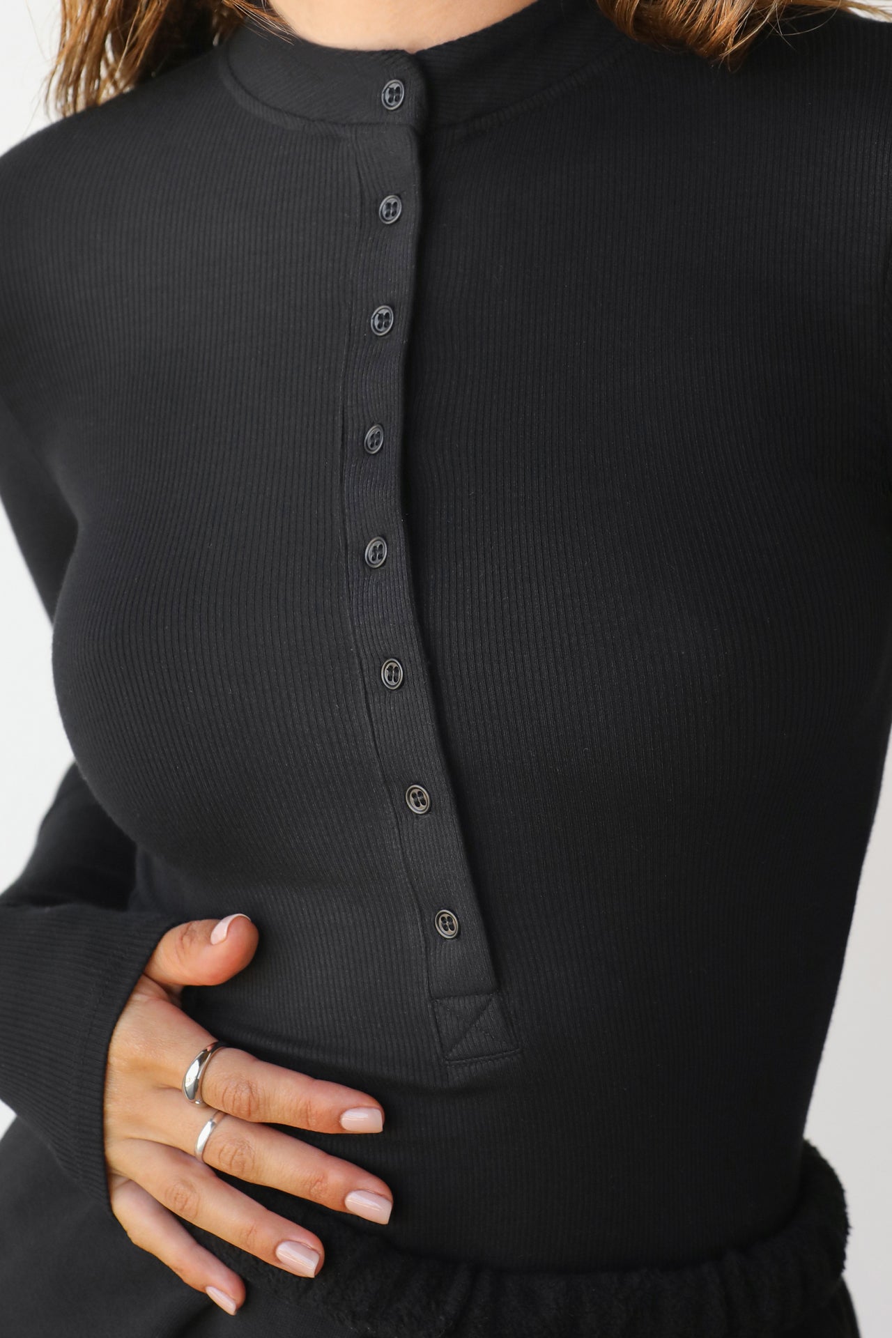 Close up detail front view of model posing in the fitted and stretchy black rib Button Down Henley long sleeve top with a high crew neckline and front buttons that reach about halfway down the shirt