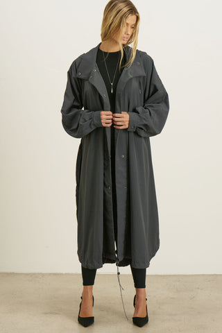 Full body front view of model posing in the jet nylon City Trench coat with oversized silhouette, structured collar, attached hood, self-tie belt and adjustable sweep